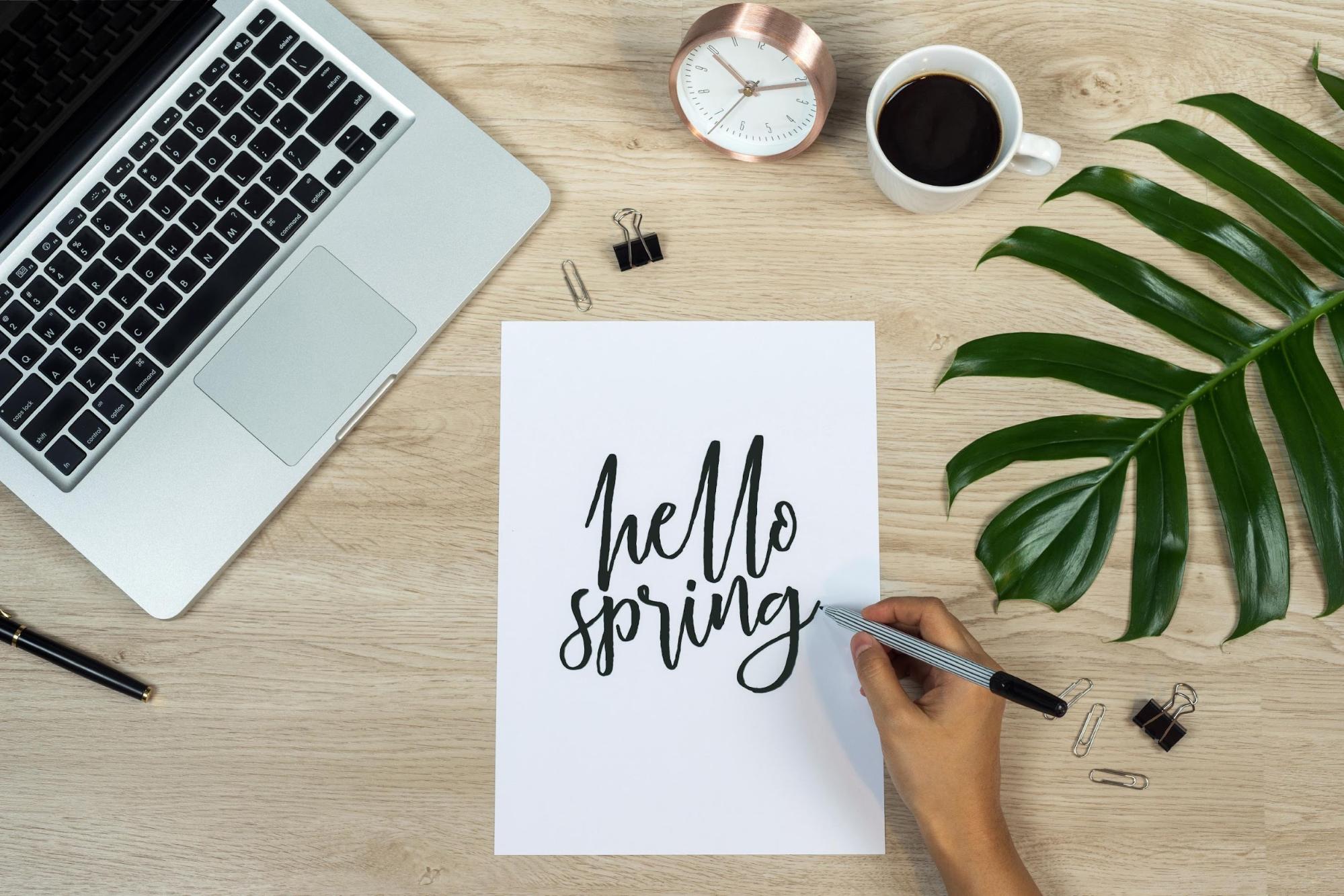 Conceptual Image of a Hand Writing “Hello Spring” on a Paper on a Desk with a Computer, Coffee, Clock, and Large Leaf