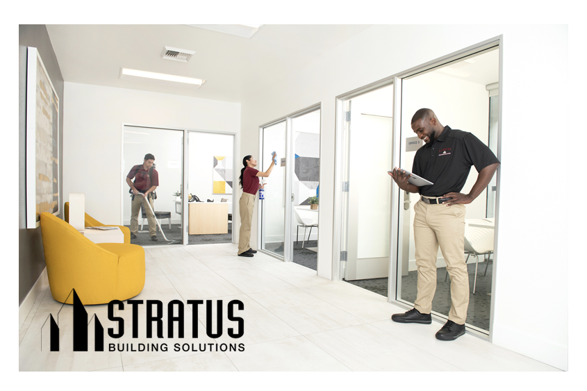 A Stratus Franchise Owner Smiles and Looks at a Tablet While Two Workers in the Background Vacuum and Wipe a Sliding Glass Door 