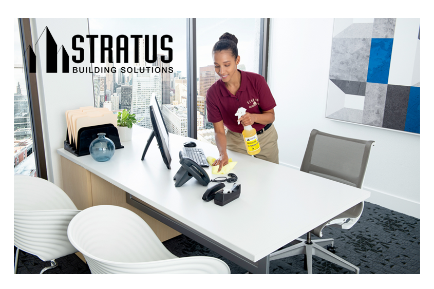 A Smiling Uniformed Stratus Cleaner Holds a Bottle of Cleaning Spray in One Hand While Wiping a Desk with the Other