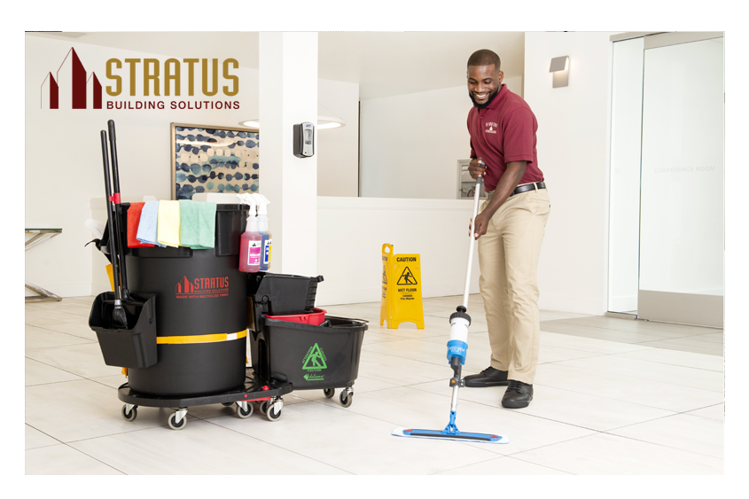 A Smiling Uniformed Stratus Cleaner with a Trash Can and Mop Bucket on Wheels with Cloths on it Next to Him Mops a Tile Floor