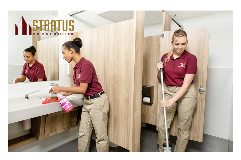 A Uniformed Stratus Employee Sprays and Wipes Down a Bathroom Counter While Another Exits a Stall While Mopping the Floor