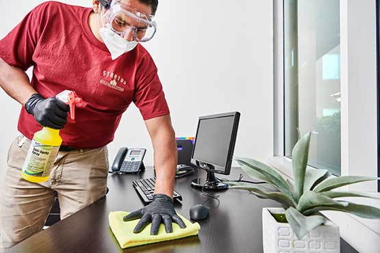 A Man in a Stratus T-Shirt, Black Gloves, and Goggles Holds a Bottle of Yellow Liquid and Wipes a Desk with a Yellow Cloth