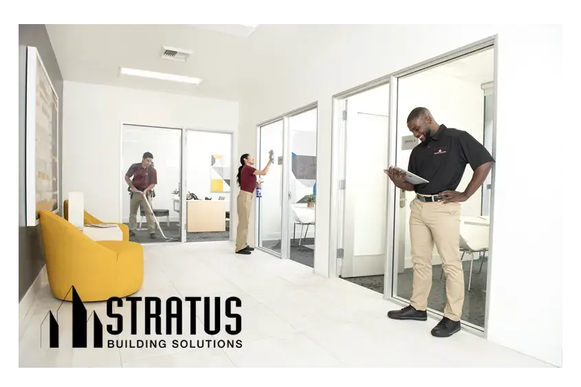 A Stratus Franchise Owner Smiles and Looks at a Tablet While Two Workers in the Background Vacuum and Wipe a Sliding Glass Door 