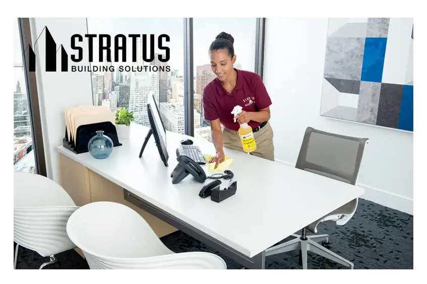 A Smiling Uniformed Stratus Cleaner Holds a Bottle of Cleaning Spray in One Hand While Wiping a Desk with the Other