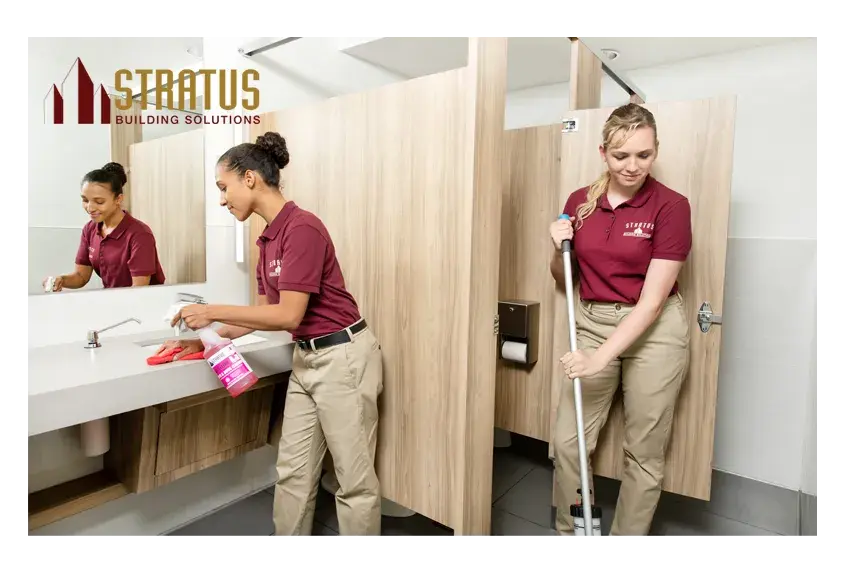 A Uniformed Stratus Employee Sprays and Wipes Down a Bathroom Counter While Another Exits a Stall While Mopping the Floor