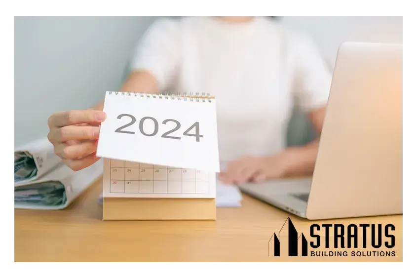 Torso of a Woman at a Table with a Laptop on it Reaching Out to Flip a Calendar with 2024 Printed on the Top Page